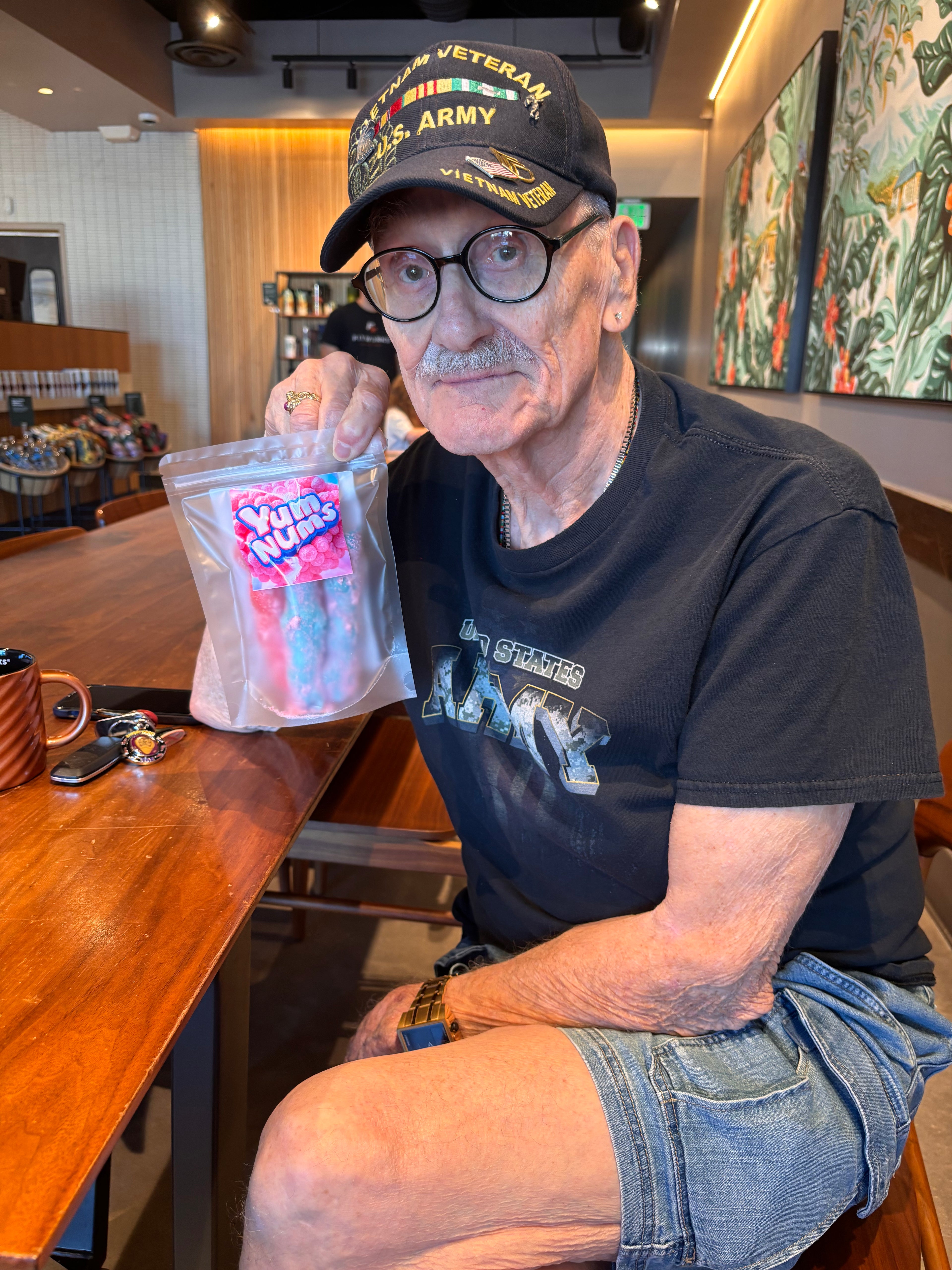 Man sitting at a starbucks holding a bag of Yumnums candy, wearing a black t-shirt with text and a cap.