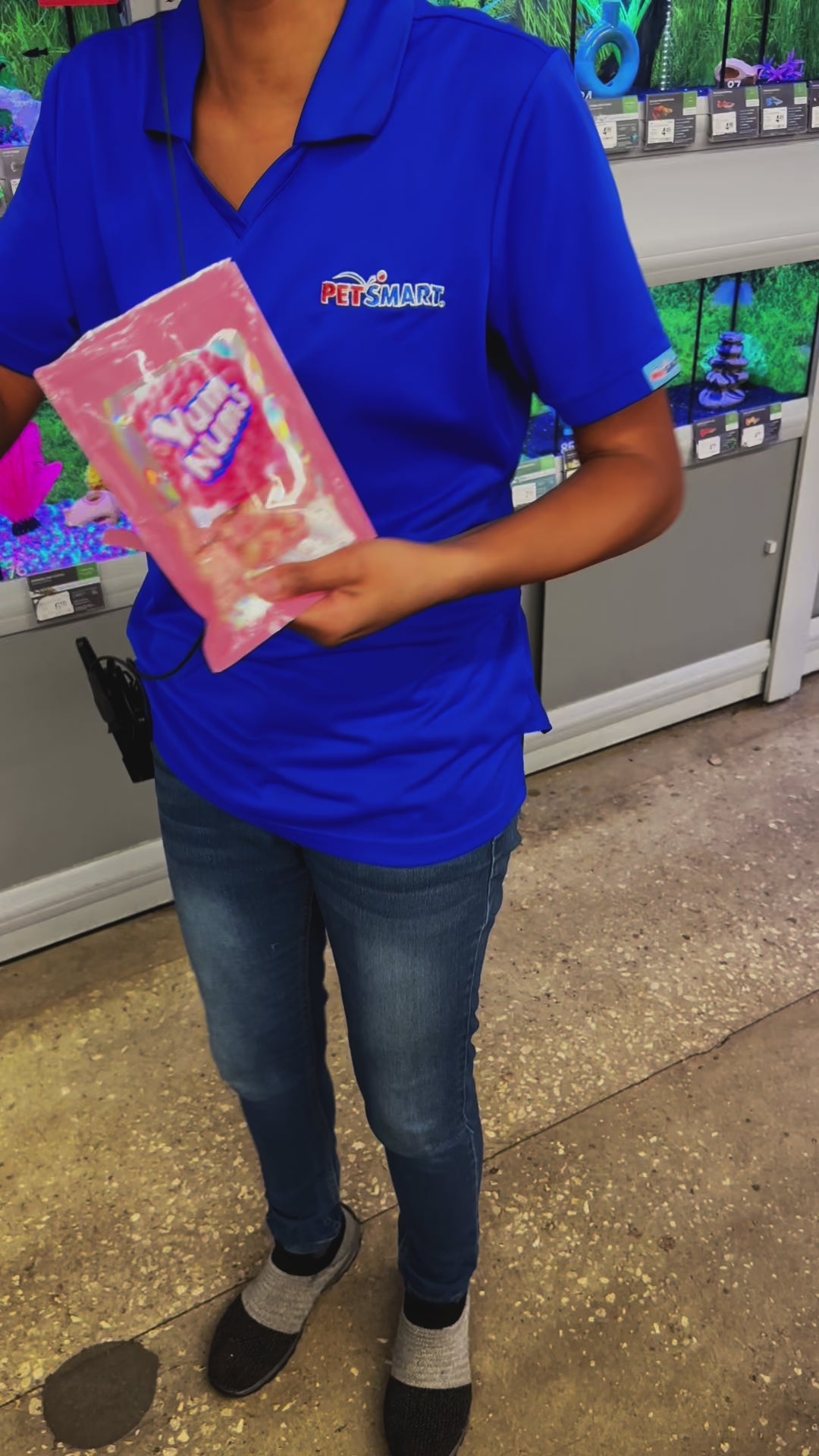 PetSmart employee in uniform holding a bag of Watermelon YumNums Freeze-Dried Candy, smiling while trying the crunchy, juicy watermelon snack for the first time in a taste-test video.